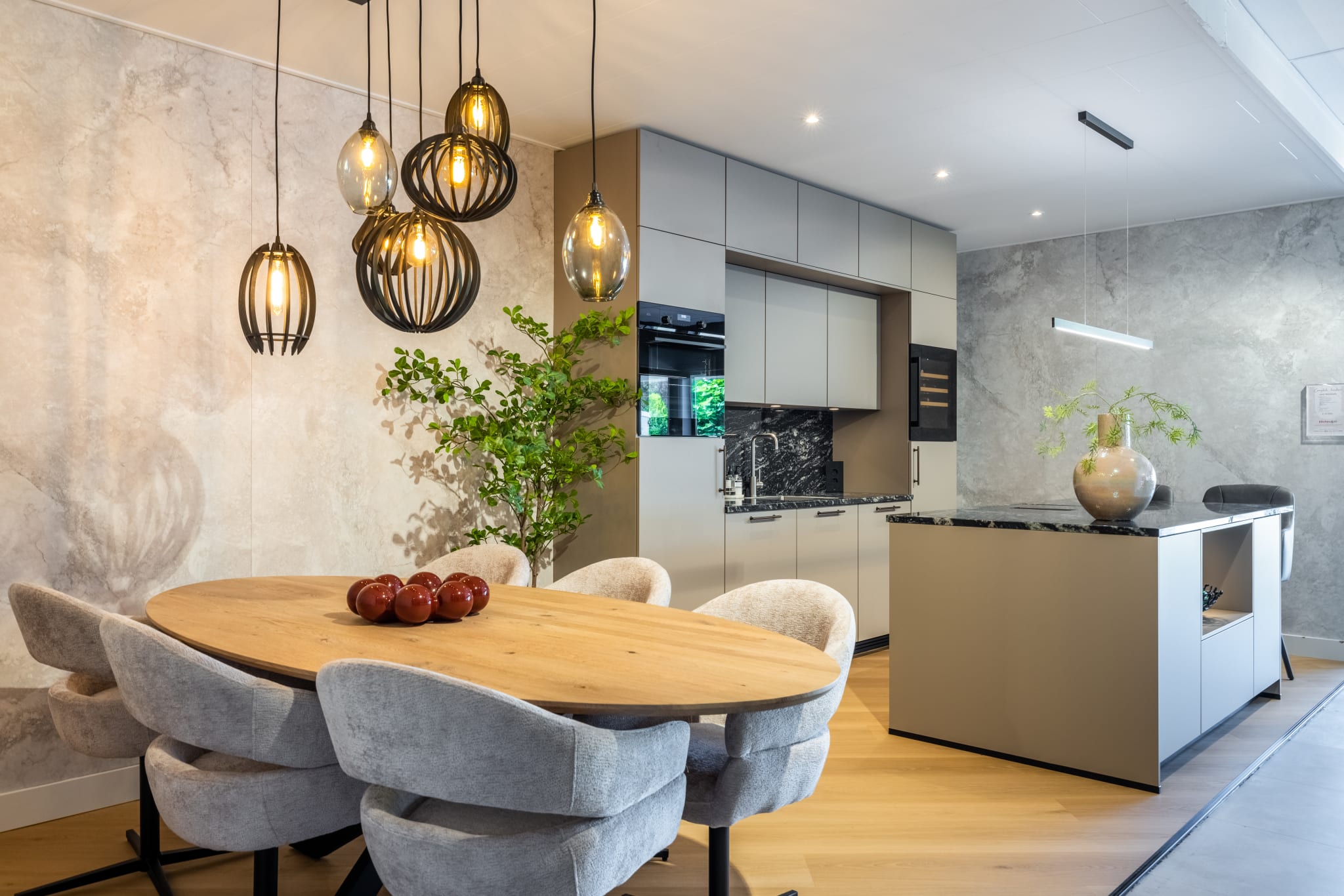 Ovale eiken eettafel met grijze stoelen vlak naast een taupe kookeiland met een donker stenen blad