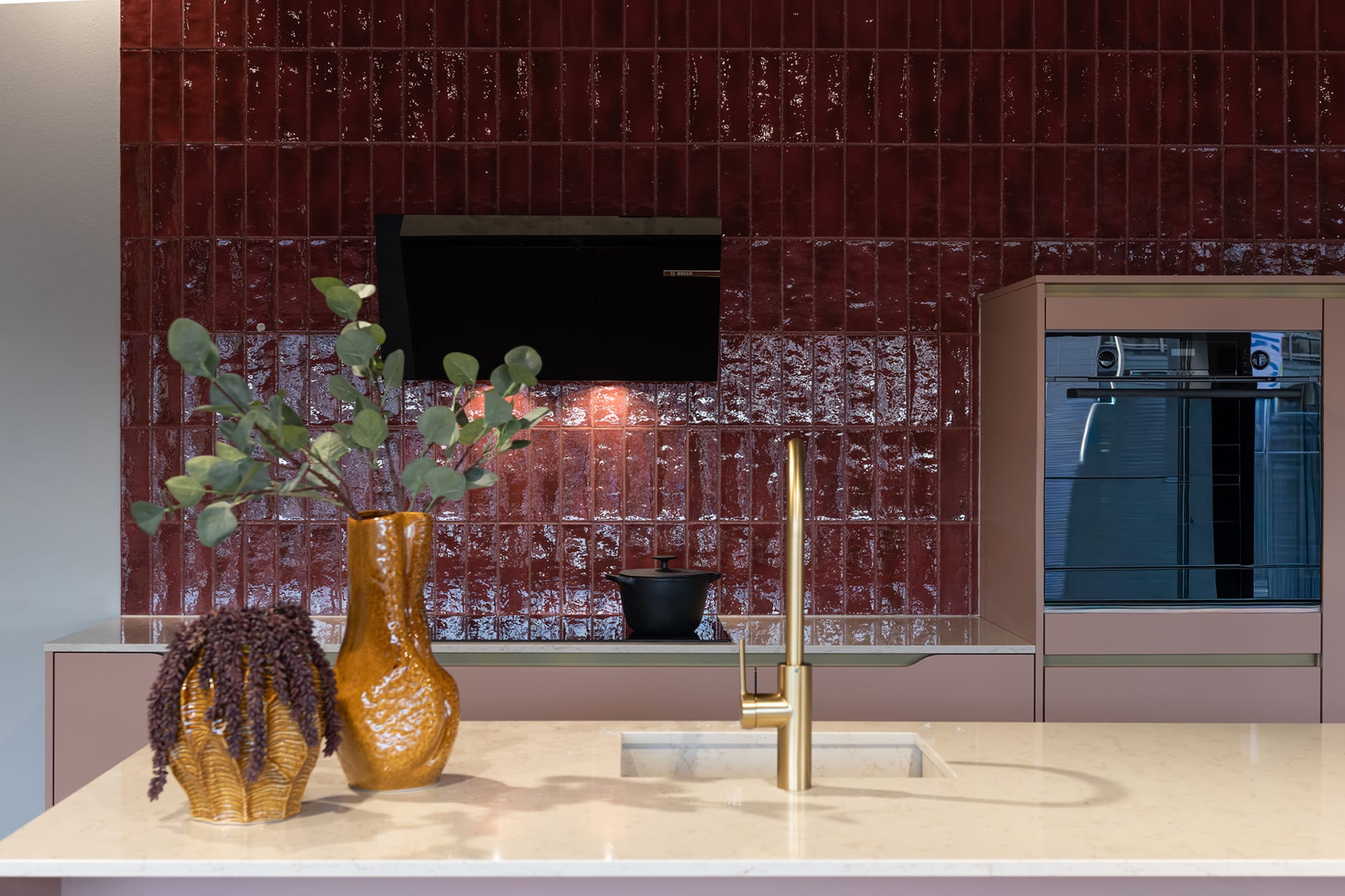 Moderne Kitchen4All eilandkeuken met beige en taupe fronten, een composiet werkblad en een opvallende rode tegelwand.