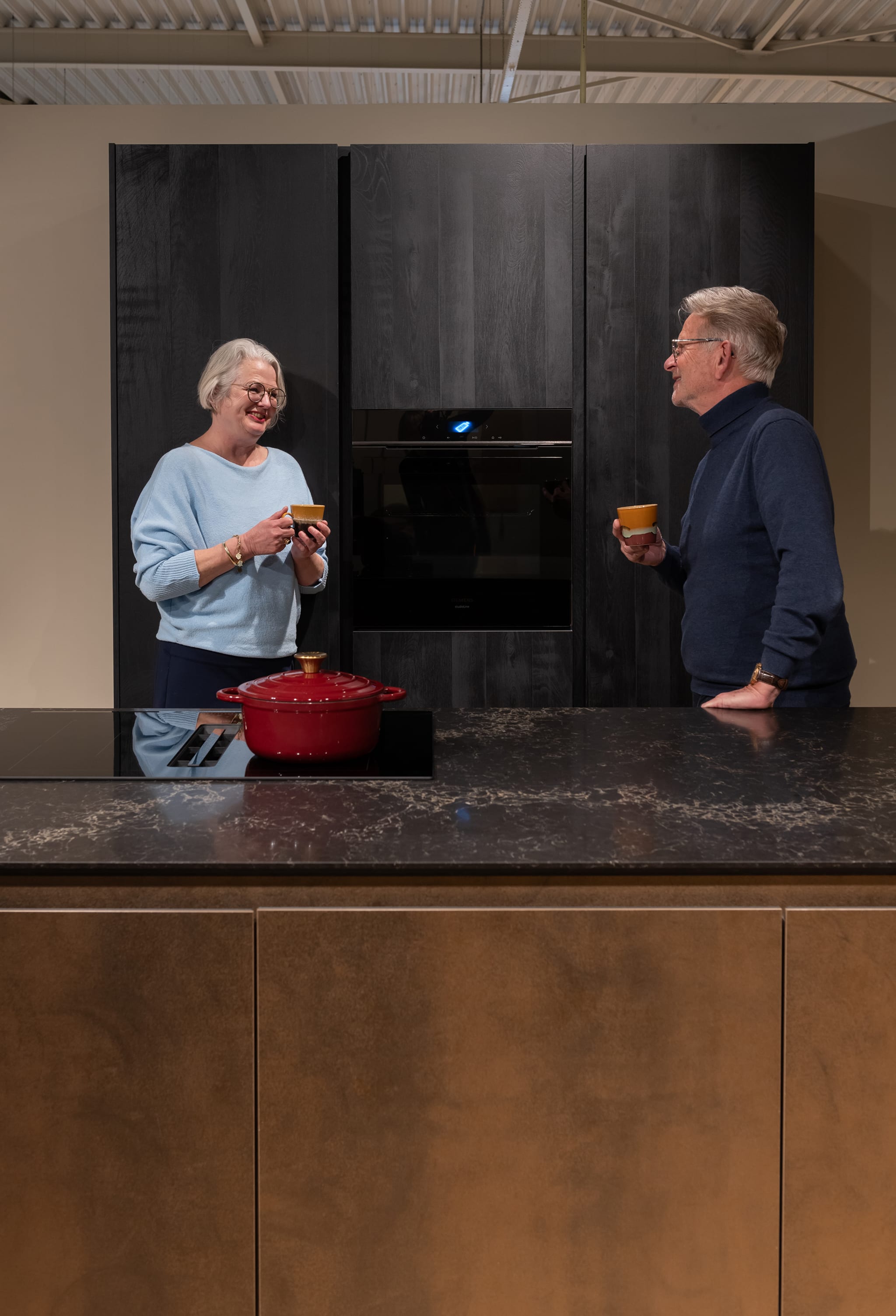Een stel geniet van koffie in een moderne chique Kitchen4All eilandkeuken met donker composiet werkblad en houten fronten.