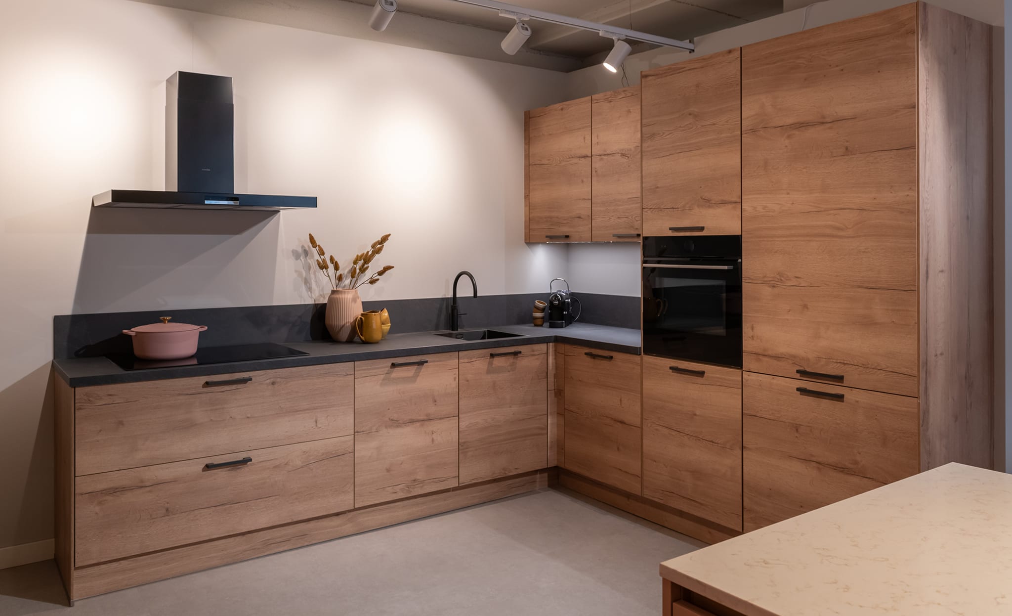 Moderne L-keuken van Kitchen4All met houtlook fronten, zwarte handgrepen en een donker composiet werkblad.