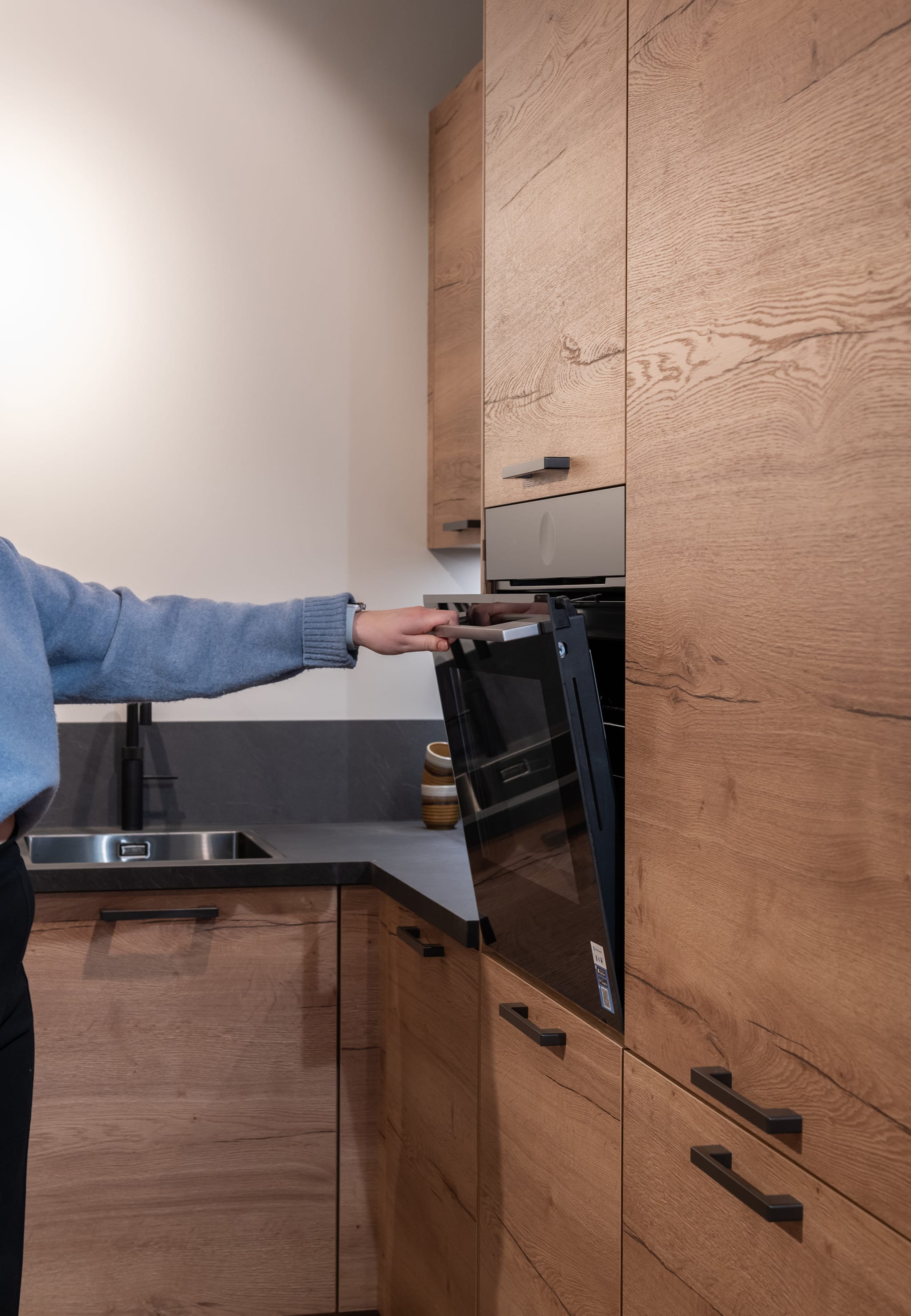 Een moderne Kitchen4All L-keuken met houtlook fronten en een donker werkblad, waarbij een oven wordt geopend.