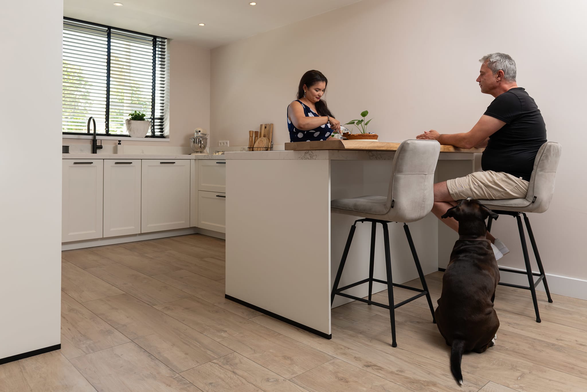 Een man zit aan het kookeiland terwijl een vrouw iets snijdt, met een hond ernaast, in een lichte keuken met houtlook vloer.
