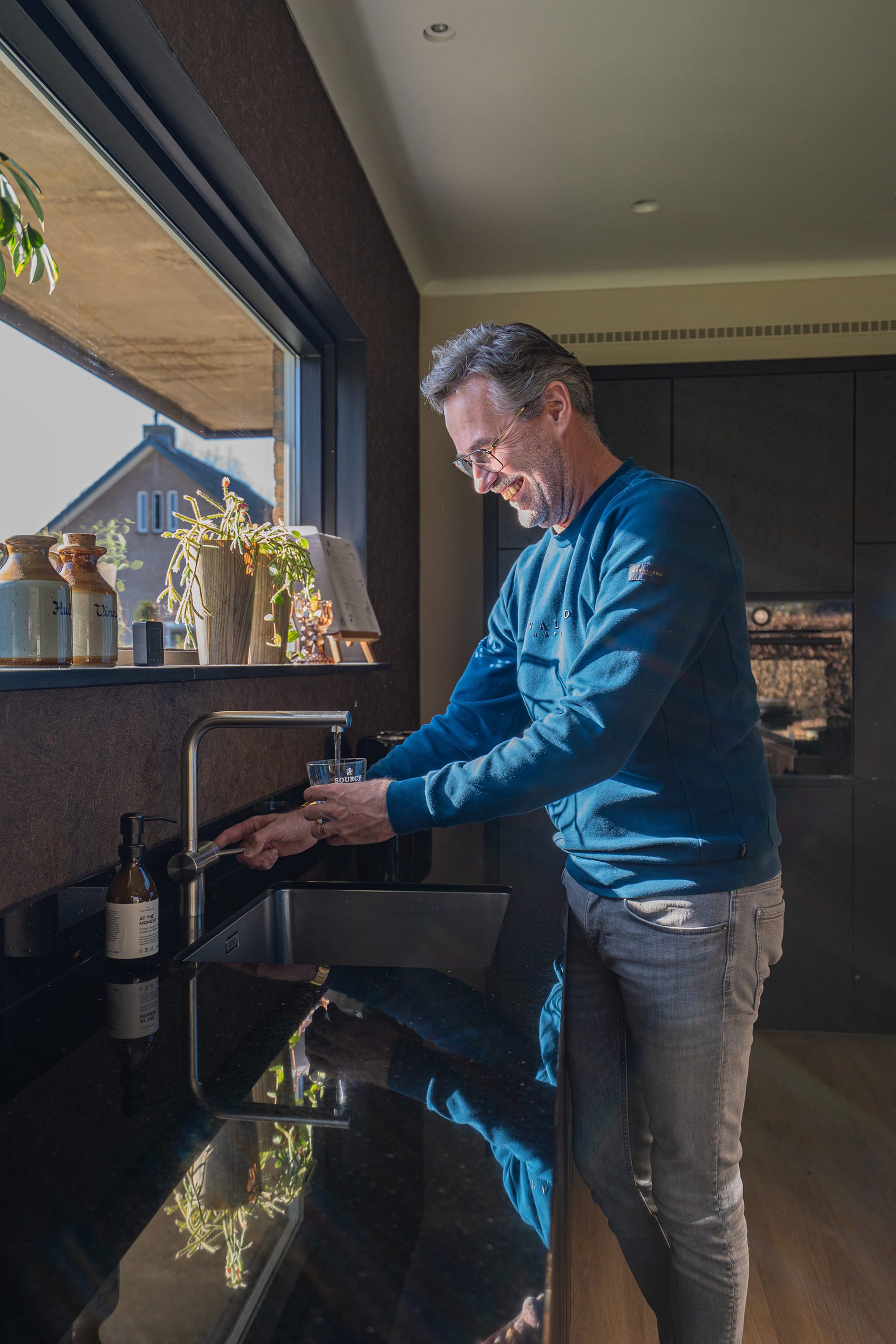 Rick tapt een glas water bij de spoelbak onder het lange raam